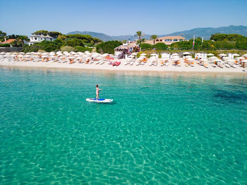 Klares türkisfarbenes Meer mit Person auf Paddleboard, weißer Sandstrand und Sonnenschirme im Hintergrund.