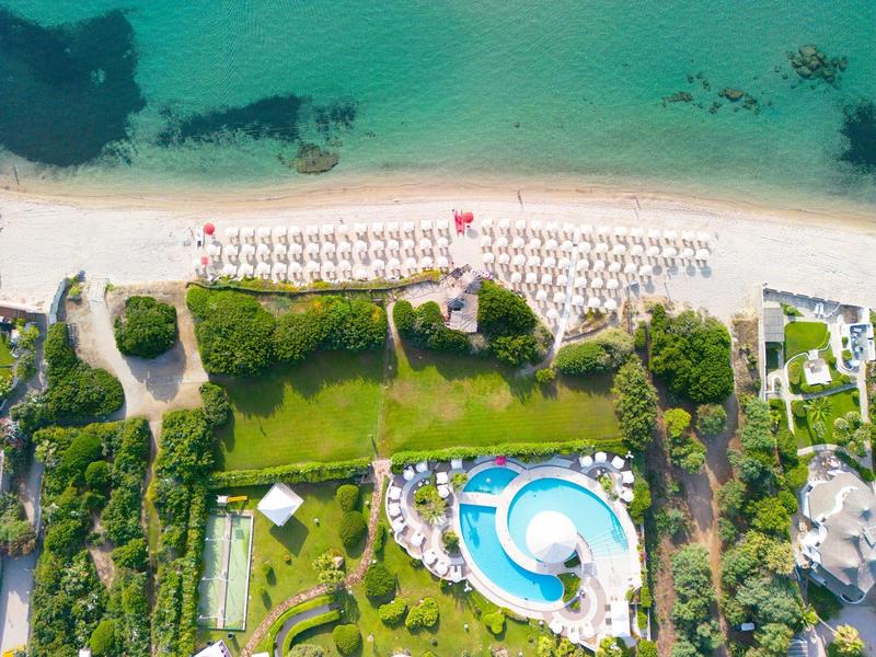 Luftaufnahme eines Strandes mit weißem Sand, blauem Meer, Sonnenschirmen und angrenzendem Poolbereich.