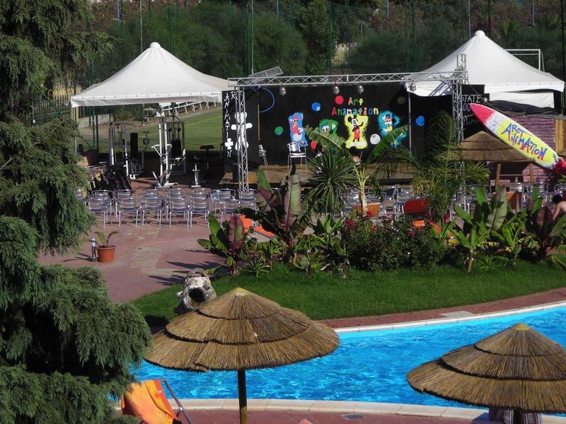 Vue d'un espace événementiel avec scène, sièges et piscine avec parasols en paille au premier plan.