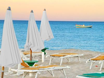 Strand mit weißen Sonnenschirmen, Liegestühlen und kleinem Boot auf ruhigem blauen Meer vor Abendhimmel.