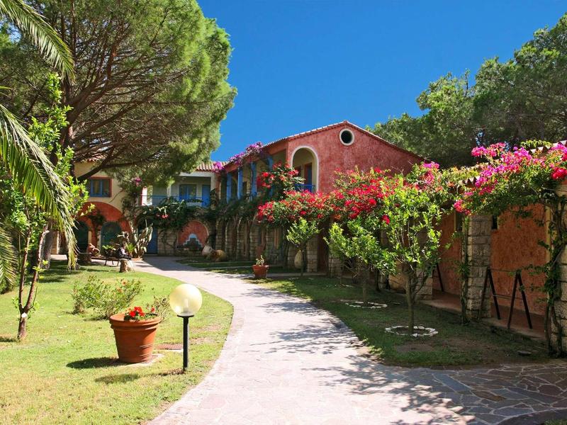 Weg vor roten Häusern mit grünen Bäumen und pinken Blumen unter klarem blauem Himmel.