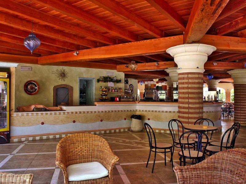 Offene Bar mit bunten Fliesen, Holzbalkendecke und Korbsesseln auf Terrakottaboden.