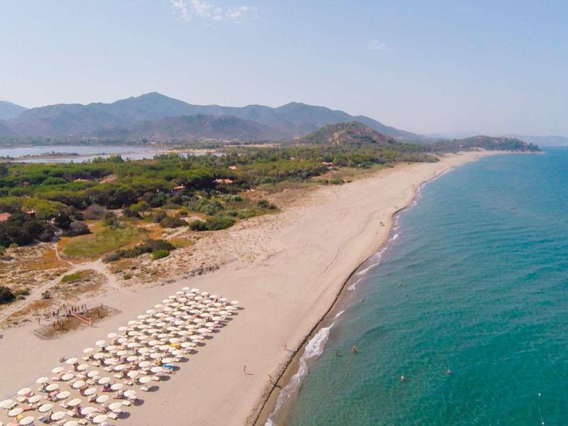 Breiter Sandstrand mit Sonnenschirmen und blauem Meer, umgeben von grüner Landschaft und Bergen.