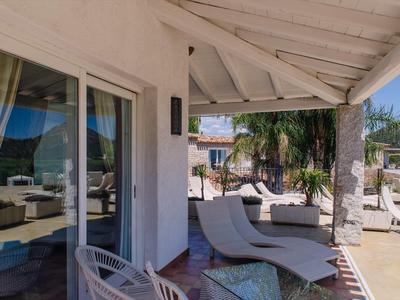 Zastřešená terasa s lehátky a skleněným stolem v hotelu s výhledem na palmy.