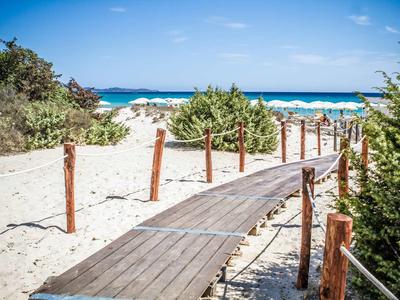 Holzsteg führt durch weißen Sand und grüne Büsche zum blauen Meer unter blauem Himmel.