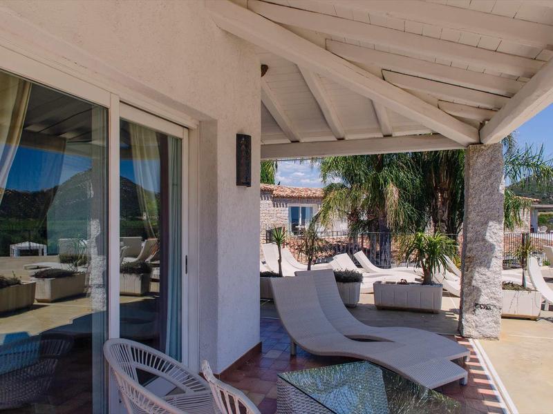 Zastřešená terasa s lehátky a skleněným stolem v hotelu s výhledem na palmy.