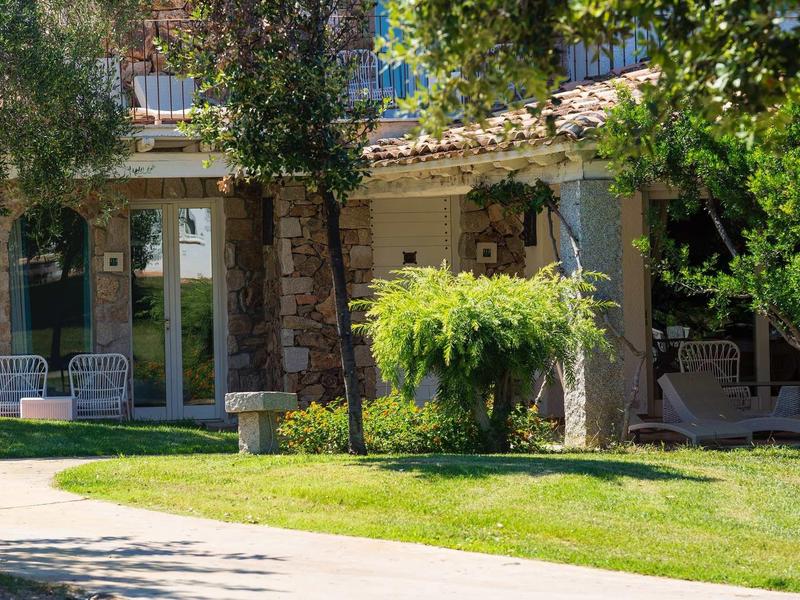 Zahrada se zelení a stromy před budovou s kamennými zdmi a židlemi.