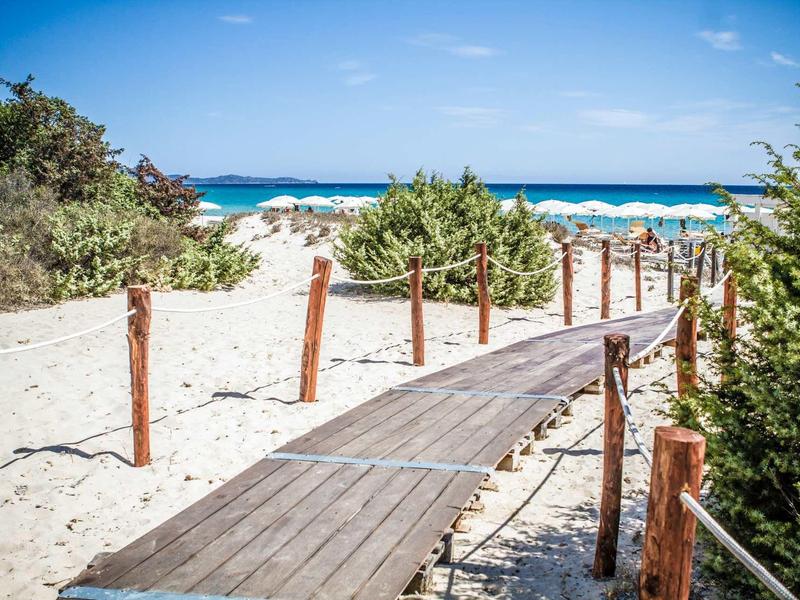 Dřevěná lávka vede přes písčité duny k čistému modrému moři pod modrou oblohou.