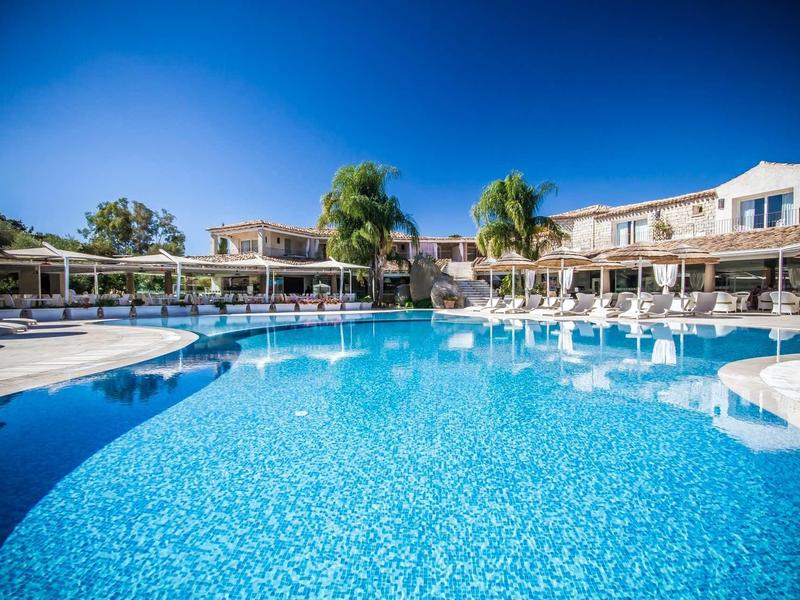 Velký venkovní bazén s lehátky a dvoupodlažními hotelovými budovami pod jasnou oblohou.