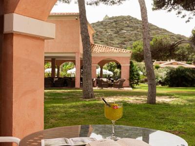 Een ronde glazen tafel met een boek, drankje en stoelen onder een boog met uitzicht op bomen en een gebouw.