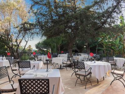 Buitentafels met witte tafelkleden en stoelen onder bomen op een zonnige dag.
