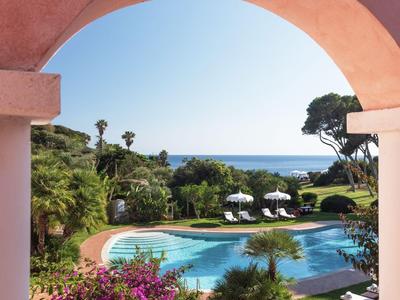 Uitzicht op een hotelzwembad met ligstoelen, palmbomen en zee aan de horizon door een roze boog.