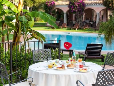 Ronde tafel met wit tafelkleed en gedekte gerechten naast een zwembad voor een huis met tuin.