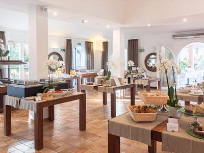 Lichte ontbijtruimte met houten tafels, diverse gerechten en grote ramen met uitzicht op groen.