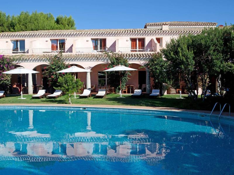 A hotel with pool, sun umbrellas, and lounge chairs in front of a two-story building with red roof tiles.