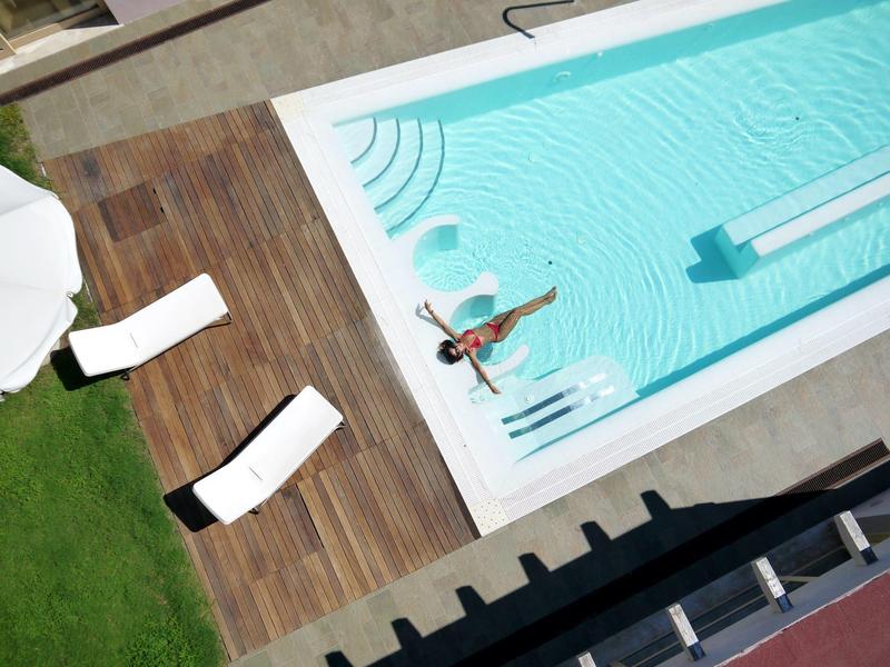 Widok z góry na basen z osobą w środku, otoczony drewnianym tarasem i leżakami.