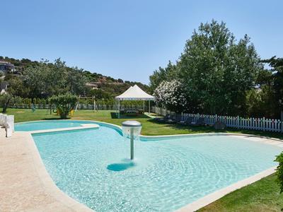 Une piscine claire avec une zone peu profonde et un parasol dans un jardin vert sous un ciel clair.