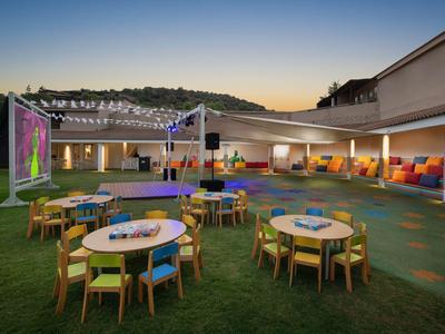 Espace extérieur moderne d'hôtel avec tables rondes, chaises et grand écran au coucher du soleil.