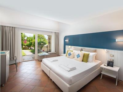 Chambre d'hôtel lumineuse avec lit double, porte terrasse et vue sur jardin avec piscine.