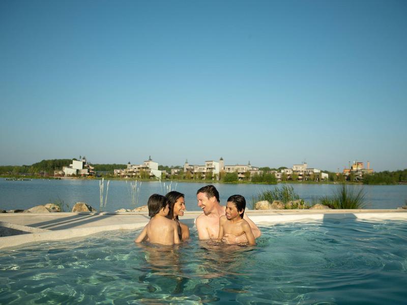 Four people relax in a hot tub overlooking a calm lake and houses on the shore.