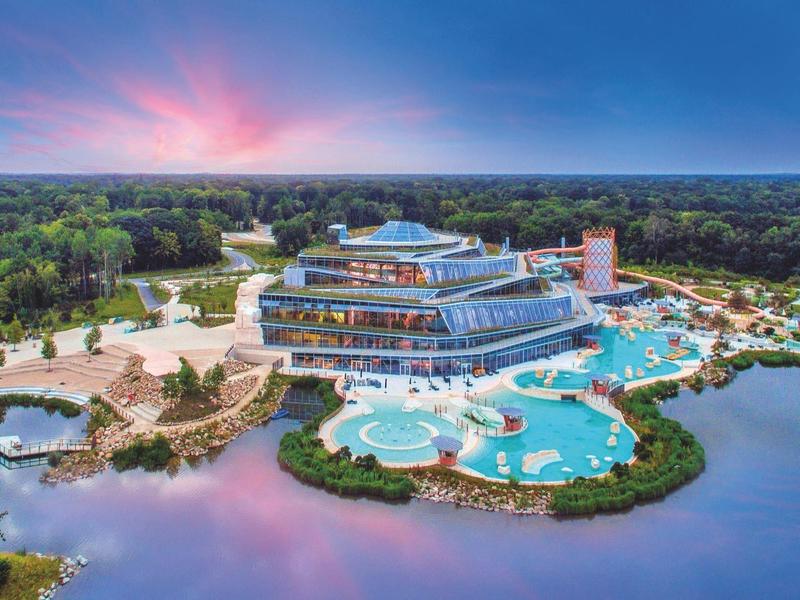 Modern hotel complex with pools surrounded by nature and water under a colorful sky.