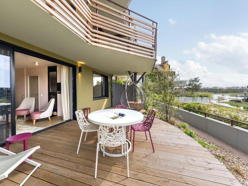 Balcony with wooden floor, round table, and colorful chairs overlooking greenery and river.