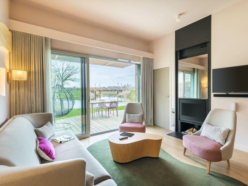 Bright hotel room with sofa, two armchairs, and view of terrace and lake through large glass doors.