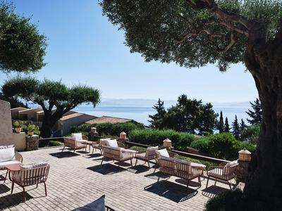 Terras met stoelen en tafels met uitzicht op zee en groene heuvels onder een heldere lucht.