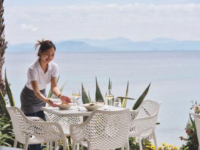 Vrouw serveert eten aan een witte tafel bij zee met blauwe lucht op de achtergrond.