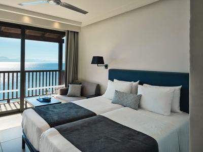 Helles Hotelzimmer mit zwei Betten, Balkon und Meerblick bei Tag.