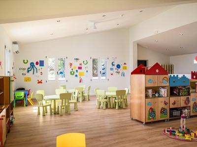 Heller Kinderspielraum mit Tischen, Stühlen, Spielzeug und bunten Wandbildern.
