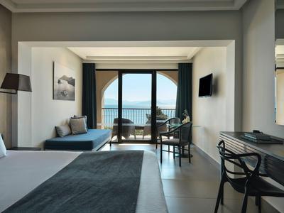 Helles Hotelzimmer mit Meerblick, Doppelbett, Sitzecke und Balkon mit Stühlen.