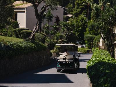 Kleiner Golfwagen fährt auf schattiger Straße zwischen grünen Büschen und Hotelgebäuden.