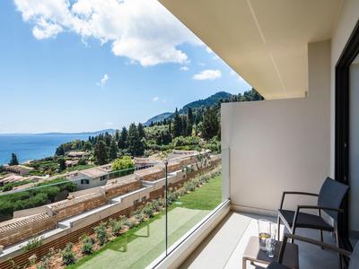 Balkon mit Glasgeländer, Stühlen und Meerblick in einer grünen, hügeligen Umgebung.