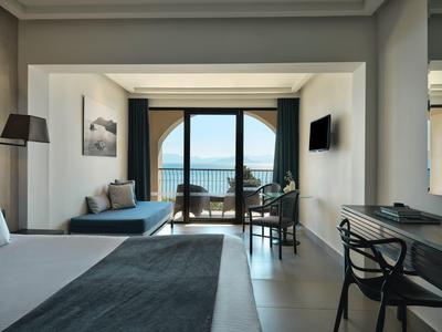 Helles Hotelzimmer mit großem Bett, Sitzecke, Schreibtisch und Balkon mit Meerblick und blauer Dekoration.