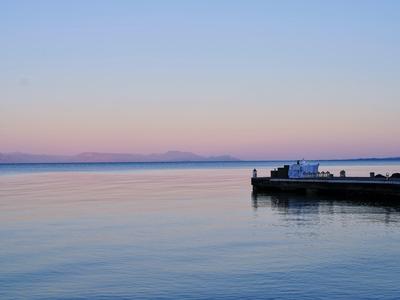 Ruhiger See mit sanften Farben bei Sonnenuntergang, rechts kleiner Steg mit Lastwagen und Menschen.