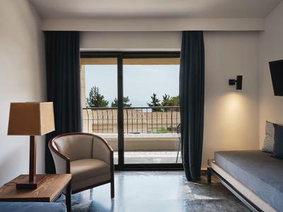 Moderner Wohnraum mit Sessel, Holztisch, Stehlampe, Sofa und großer Fensterfront mit Meerblick.