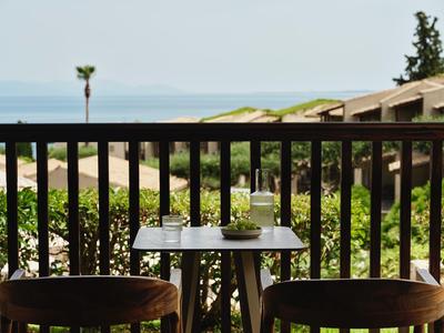 Balkontisch mit zwei Stühlen, Wasserflasche und Gläsern, Blick auf Meer und grüne Landschaft.