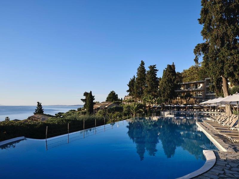 Infinity-Pool mit spiegelglattem Wasser vor blauen Himmel, umgeben von Bäumen und Liegestühlen.