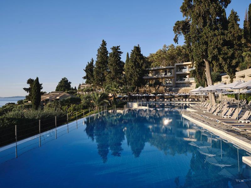 Großer blauer Pool mit Sonnenliegen am Rand und Bäumen vor einem mehrstöckigen Gebäude.