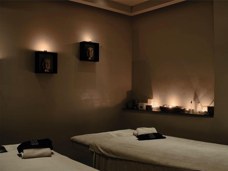 Dunkler Raum mit zwei Massageliegen und gedämpfter Beleuchtung an Wänden und Regalen.