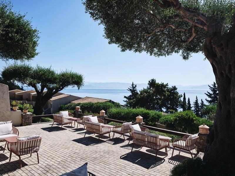 Terrasse mit Holzstühlen und Tischen, Blätter und Meerblick bei klarem Himmel und Sonnenschein.