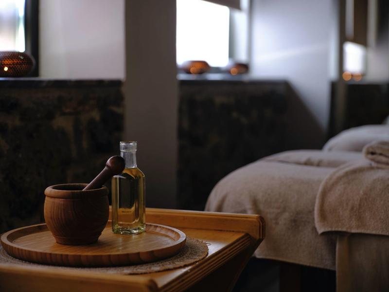 Ein Holztisch mit einer Runde Platte, einem Mörser und einem Fläschchen Öl neben einer Massageliege.