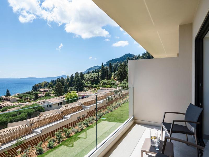 Balkon mit Glasgeländer, Stühlen und Meerblick in einer grünen, hügeligen Umgebung.