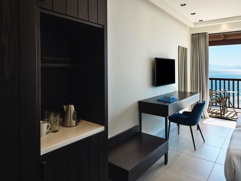 Moderner Hotelraum mit Schreibtisch, Fernseher, Stuhl, Teekocher und Balkon mit Meerblick.
