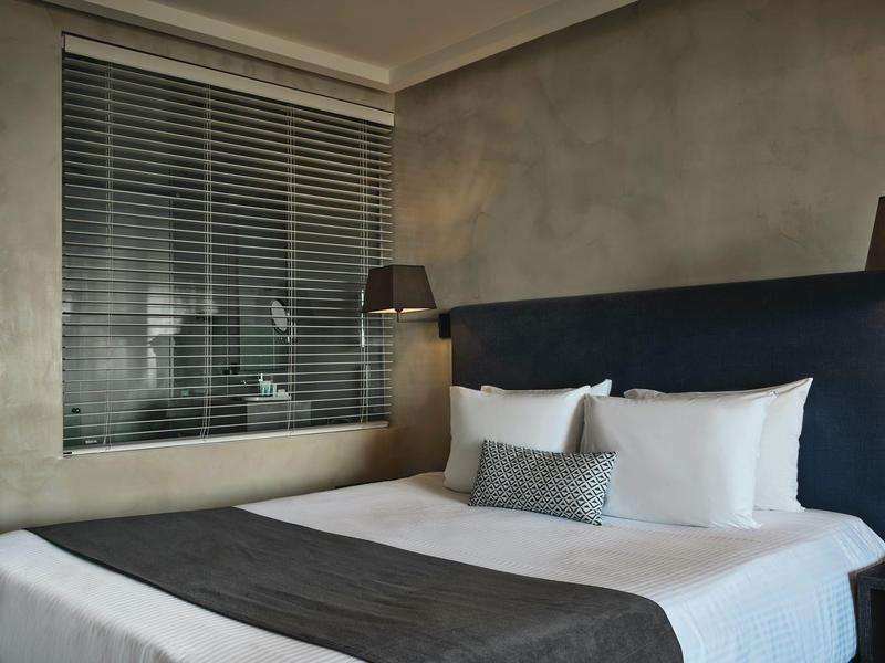 Modernes Schlafzimmer mit grauen Wänden, großem Bett, weißen Kissen und Blick durch Jalousien.