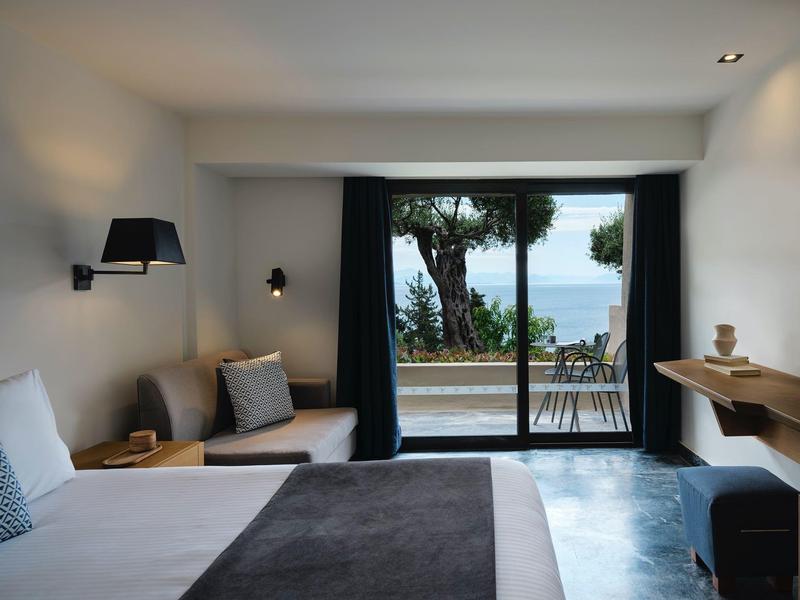 Modernes Schlafzimmer mit großem Bett, Sitzbank, Schreibtisch und Meerblick durch Glaswand.