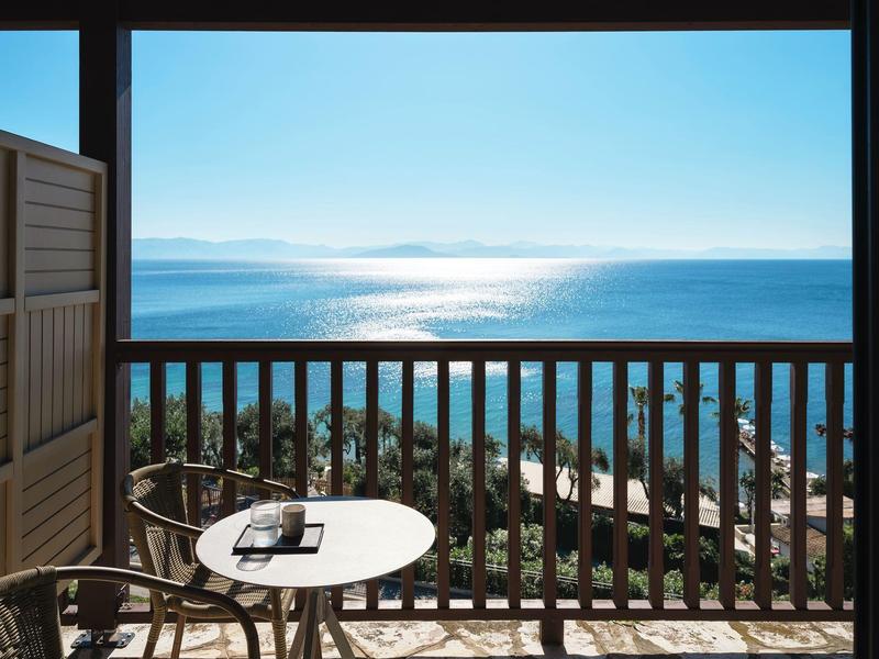 Balkon mit rundem Tisch und Stuhl, Blick auf Meer und bewaldete Küste bei klarem Himmel.