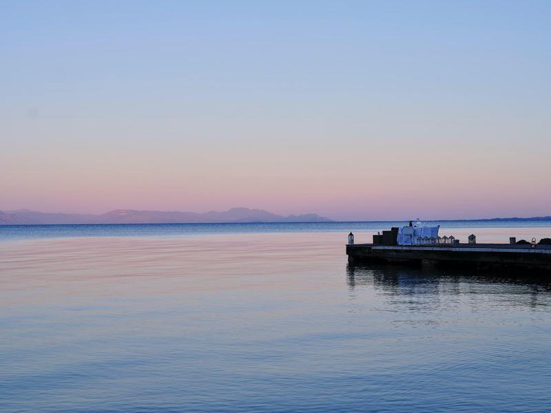 Ruhiger See mit sanften Farben bei Sonnenuntergang, rechts kleiner Steg mit Lastwagen und Menschen.