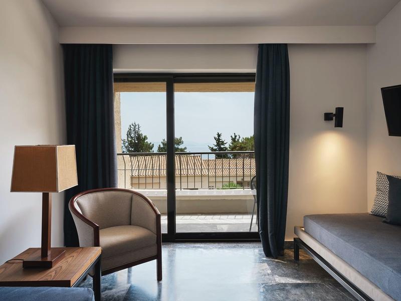 Moderner Wohnraum mit Sessel, Holztisch, Stehlampe, Sofa und großer Fensterfront mit Meerblick.